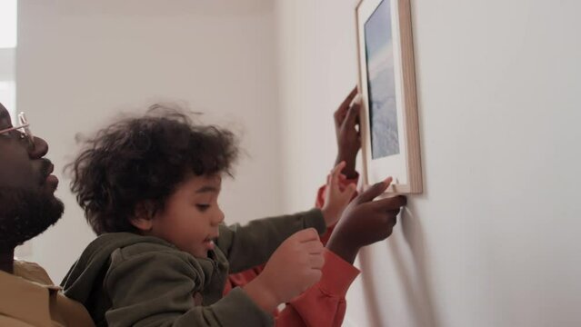 Little African American boy hanging framed picture on wall with assistance of parents while moving in new house with family