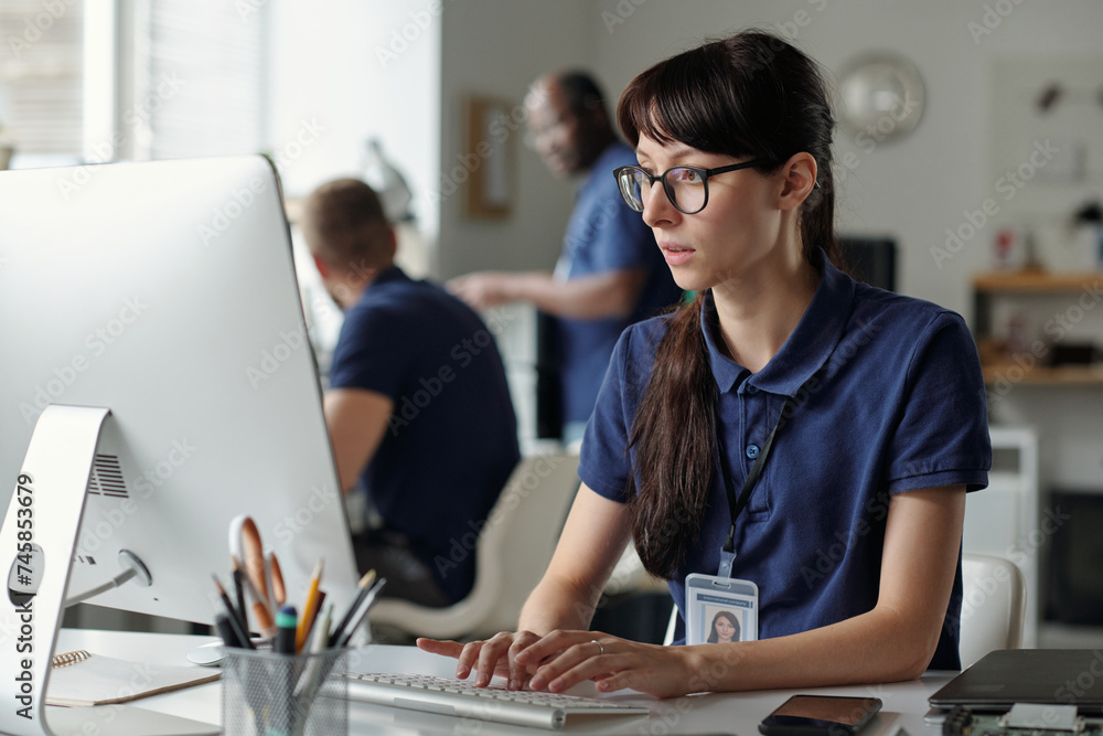 Poster young female worker of repair service center typing on computer keyboard and looking at screen while
