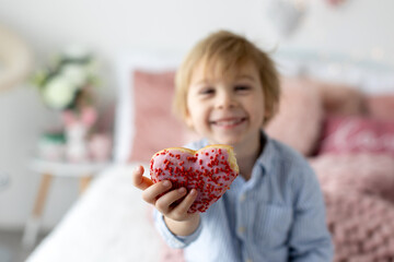 Cute little blond child, preschool boy, eating pink donut in the shape of heart, made for Valentine