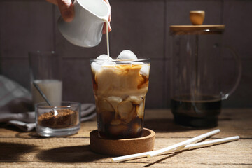 Woman pouring milk into glass with refreshing iced coffee at wooden table, closeup