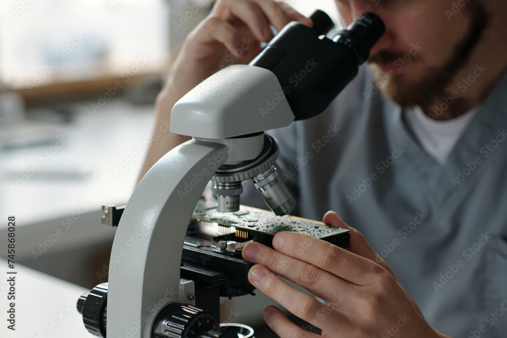 Poster close-up of young repairman looking at motherboard details through microscope lens while sitting in 