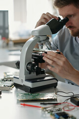 Microscope and hand of young technician of repair service center looking at motherboard details while sitting in front of camera in laboratory