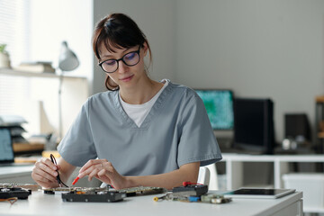 Professional technician in uniform and eyeglasses sitting by workplace in repair service center and checking details of computer processor