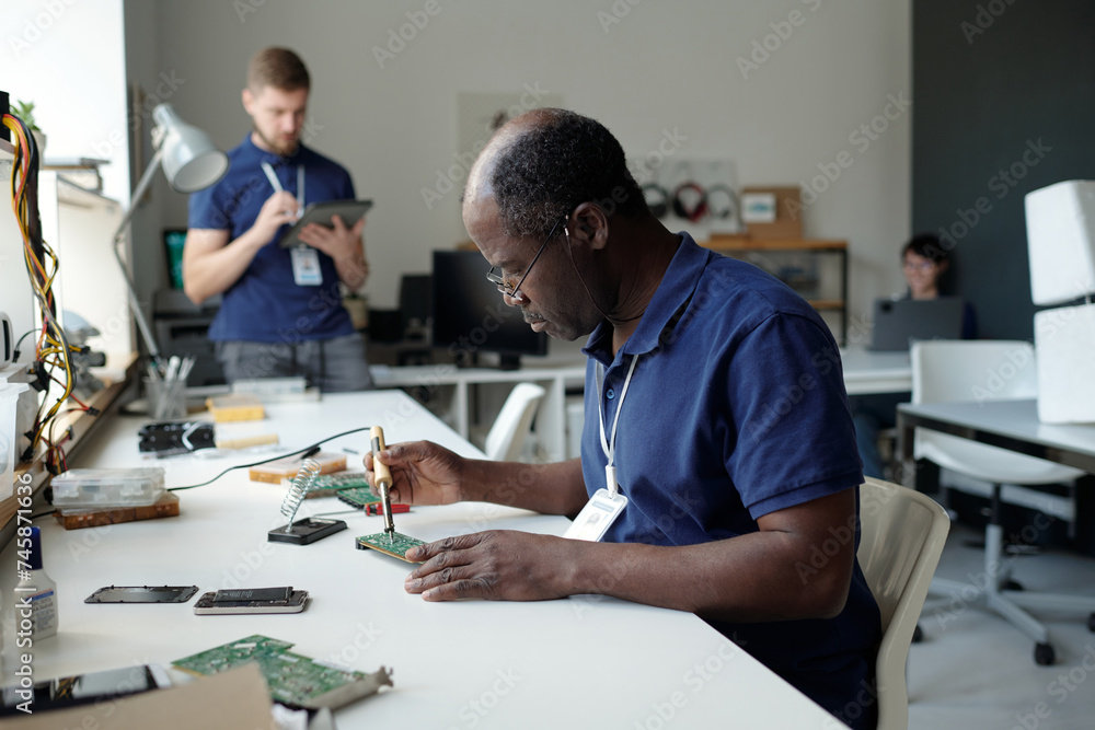 Poster experienced male technician bending over desk in coworking space and checking computer motherboard w