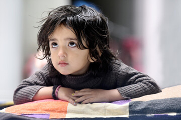 Naklejka premium 3 years plus cute Asian Indian baby girl in open hair wearing brown high-necked sweater looking up, giving innocent expressions while lying on the floor and keeping her hands on the pillow