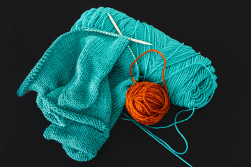 Teal coloured knitted item on circular needles and ball of rust coloured wool displayed with skein of teal yarn viewed from above on a black background
