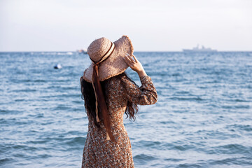 Slim woman in summer dress and straw hat standing on a beach. Dreaming girl, romantic vacation at the sea resort
