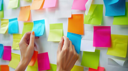 Fototapeta premium Close-up of hands young marketing woman engaged in a brainstorming session, using colorful sticky notes on a white wall to organize their Ideas, Goals, Objectives, Plans and Strategies in office.