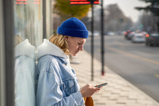 Stylish hipster, generation Z representative, sitting at bus stop, waiting for transport, messaging in smartphone, surfing in internet, scrolling web pages, photos in social networks, reading news