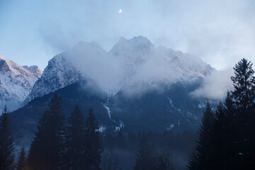 Evening scene of a mountain range with the moon in the sky, light fog, mountains in the background.