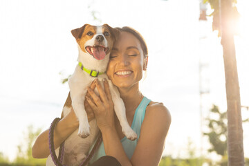 A happy girl owner with closed eyes holds her pet dog breed Jack Russell Terrier in arms lovingly in bliss, standing outdoors in the summer sunlight. The concept of true and sincere friendship between