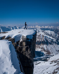 Man walking along steep mountain cliff in snow in winter, looking at mountain panorama on a sunny blue sky day. - 745923857