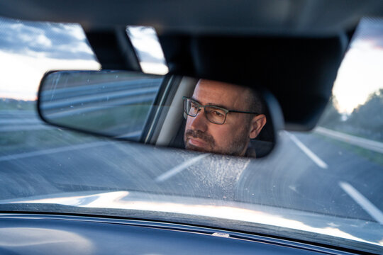 reflection in car mirror of man driving in his forties. Caucasian with glasses, bald and handsome. Through the front window we can see the lines of the road.