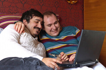 Caucasian gay couple in bed looking at laptop smiling together, side view, horizontal image with copy space. Concept of homosexual relationship