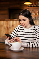 Young beautiful woman drinking coffee and looking at smartphone while sitting at cafe. Student girl using mobile phone.