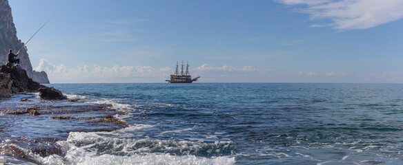 Panoramic view of the rocky coast, the silhouette of a fisherman with a fishing rod and a schooner sailing along the Mediterranean Sea in the city of Alanya, Turkey