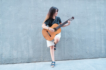 full view of a woman playing the guitar leaning against a neutral grey wall looking to the side.