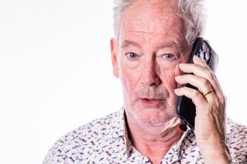 This is an image of an elderly Caucasian man with grey hair, speaking on a black smartphone with a surprised expression. He is wearing a white shirt with a floral pattern, a wedding ring, and has a