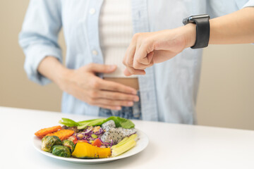 Intermittent fasting, diet healthy asian young woman dietary, having stomach ache, temptation hungry of brunch food in morning on table at home, waiting time to eat salad but not yet time first meal.
