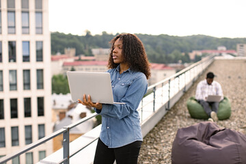 Successful business woman dressed in denim shirt and pants standing on roof top outdoors and typing on modern laptop, African American colleague works sitting in bag chair in background.