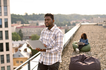 Attractive African American male freelancer using portable computer for remote work at roof top of office, during break. Blurred background of businesswoman colleague works sitting in bag chair.