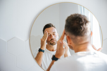 Skin care routine, young man massages his face with a cream in front of the mirror one morning in the bathroom illuminated by sunlight - 745992225