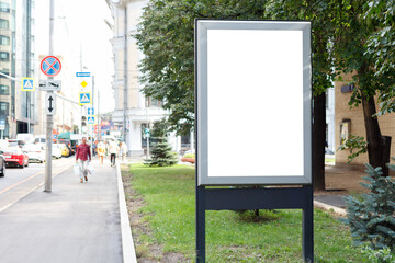 Gray empty billboard on a green lawn along a crowded street in the city in the summer. Mock-up.