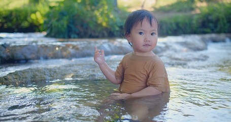 Exuberant toddler splashing in a shallow stream, laughter sparkling as sunlight filters through green foliage in a blissful outdoor scene