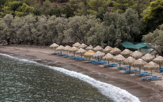 lounge chairs with straw umbrellas on empty volcanic beach in greece (european travel Mediterranean) empty loungers on shore, methana