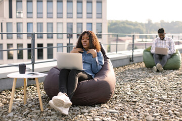 Happy African American female person in casual clothing gesture at laptop camera on flat roof of building, while her male colleague working in blurred background.