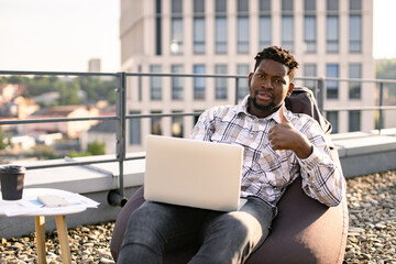 Successful African business man dressed in checkered shirt and pants sitting on beanbag on rooftop outdoors and typing on modern laptop in background of urban city view. - obrazy, fototapety, plakaty