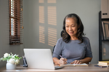 Senior asian woman smiling while working on a laptop at home. Lifelong learning and remote working concept.