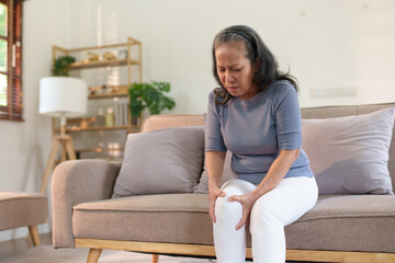 Senior asian woman experiencing knee pain sitting on sofa at home. Health issue and senior care concept.