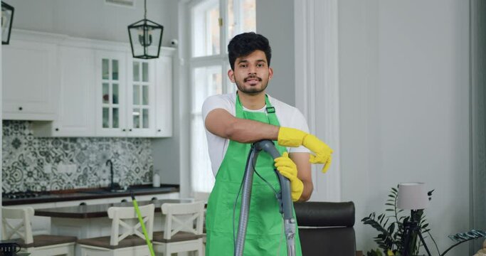 Professional man worker of cleaning service leaning on vacuum cleaner and looking at camera while cleaning up modern house