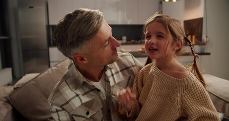 Close-up of a cunning little brown-haired girl in a cream sweater talking while her middle-aged dad with gray hair holds her in his arms while sitting on the sofa in a modern apartment