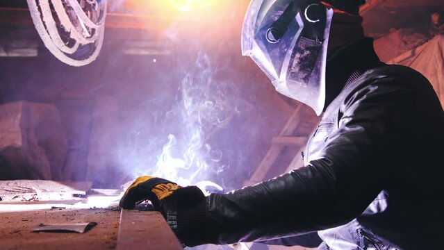 A welder in a protective mask and leather jacket is welding in his atmospheric workshop. Low key. Real time