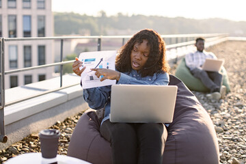 Focused African adult pointing at paper infographics during online meeting held outdoors via modern...