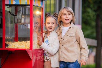 school-age boy and girl look at a popcorn machine in city park.