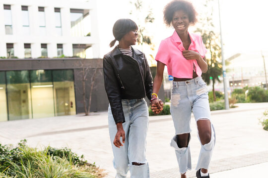 Two African American sisters walk hand in hand, laughing and enjoying each other's company in an urban setting at golden hour