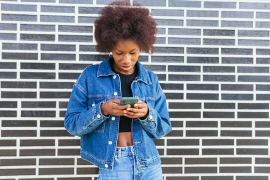 A young African American woman focuses intently on her smartphone, standing before a striking black and white brick wall
