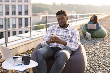 Close up view of charming African man holding phone while sitting in beanbag near coffee table with...