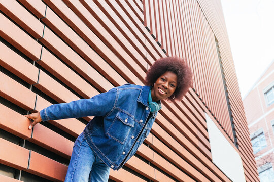 Low angle of cheerful young woman with an infectious smile leans playfully against a modern building exterior, exuding casual urban style