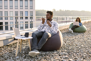 Fototapeta na wymiar Young businessman wearing checkered shirt while holding smartphone and recording audio message, sitting in pouf chair outdoors while female colleague working on blurred background.