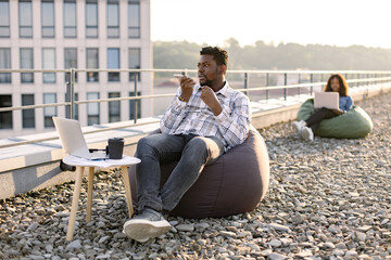 Young businessman wearing checkered shirt while holding smartphone and recording audio message, sitting in pouf chair outdoors while female colleague working on blurred background.