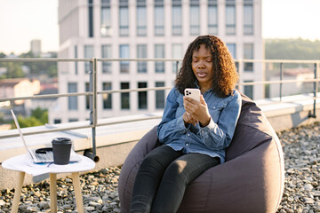 Cheerful African American person with smartphone resting in pouf chair on roof terrace during coffee break on mid day. Happy businesswoman in casual wear texting messages via mobile in open air.