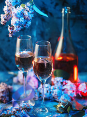 A spring celebration, a composition with champagne glasses on the table and blooming lilac branches on a blue background. Romantic Holiday Greeting Card