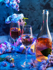 A spring celebration, a composition with champagne glasses on the table and blooming lilac branches on a blue background. Romantic Holiday Greeting Card