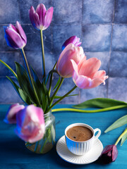 white ceramic cup of fragrant coffee on the table, a bouquet of pink tulips in a vase. blue background