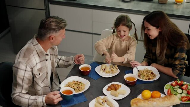Top view of a happy family having dinner at the dining table in the evening in a modern room. Happy little girl uses a table knife to eat while communicating with her parents a middle-aged man with
