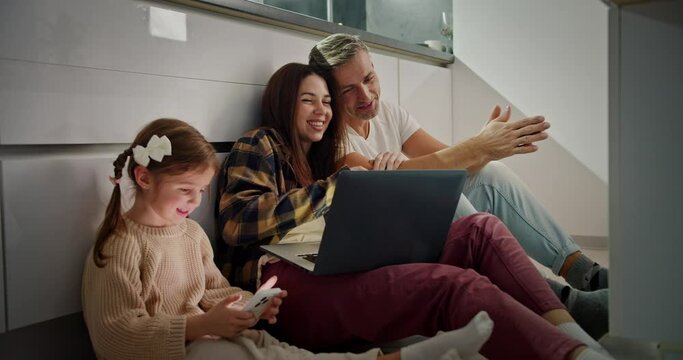 A Little Girl Is Doing Something On The Phone While Her Parents Are Sitting Next To Her And Looking At Something On The Laptop And Thinking About Their Plans. A Little Girl Is Having Fun With Her